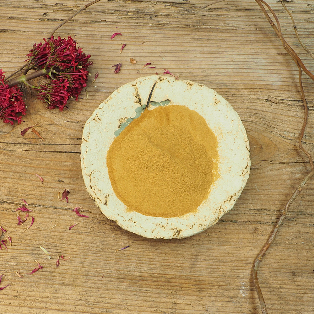 Ein rustikales Holztisch-Setting mit einer handgefertigten, leicht antiken Schale, die mit goldgelbem Parakresse Pulver-Extrakt gefüllt ist. Daneben liegt eine getrocknete Blume mit tiefroten Blüten. Das Parakresse Pulver-Extrakt ist ein naturkosmetischer Rohstoff, bekannt für seine hautpflegenden und revitalisierenden Eigenschaften. Ideal für die Herstellung von natürlichen Kosmetikprodukten.